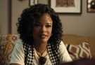 "A young woman with curly hair sits on a patterned couch in a cozy living room, wearing a checkered vest and white shirt, with framed art and cushions in the background, engaged in conversation."