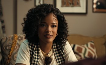 "A young woman with curly hair sits on a patterned couch in a cozy living room, wearing a checkered vest and white shirt, with framed art and cushions in the background, engaged in conversation."