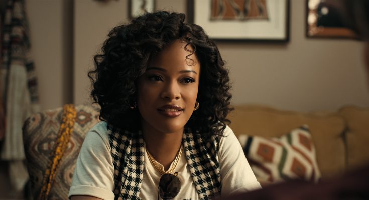 "A young woman with curly hair sits on a patterned couch in a cozy living room, wearing a checkered vest and white shirt, with framed art and cushions in the background, engaged in conversation."