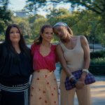 "Three women standing close together outdoors on a sunny day in a lush park, smiling and posing for a photo with trees, greenery, and a body of water in the blurred background."