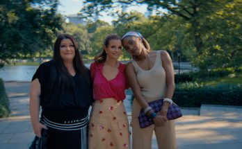 "Three women standing close together outdoors on a sunny day in a lush park, smiling and posing for a photo with trees, greenery, and a body of water in the blurred background."