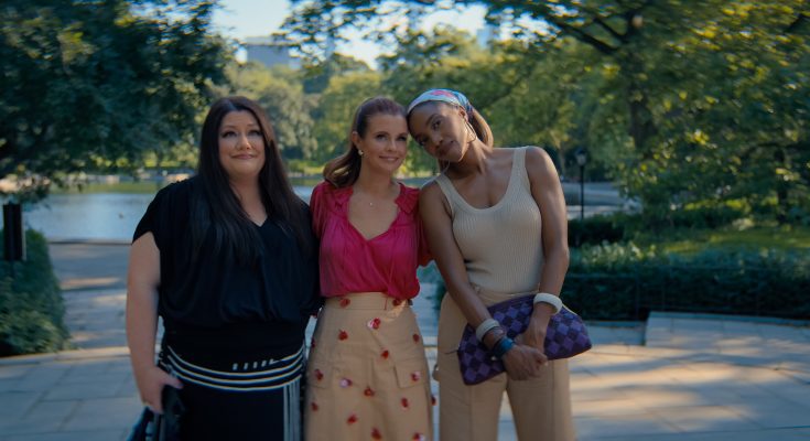 "Three women standing close together outdoors on a sunny day in a lush park, smiling and posing for a photo with trees, greenery, and a body of water in the blurred background."