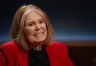 "Smiling elderly woman with shoulder-length light hair, wearing a red blazer and black shirt, sitting indoors in a dimly lit interview or studio setting with a dark, softly illuminated background."