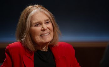 "Smiling elderly woman with shoulder-length light hair, wearing a red blazer and black shirt, sitting indoors in a dimly lit interview or studio setting with a dark, softly illuminated background."