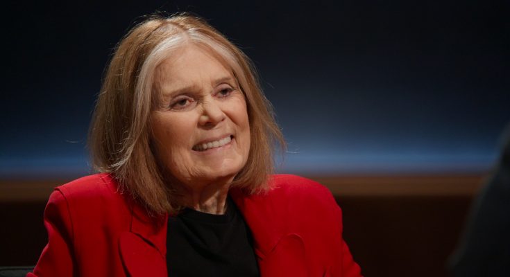 "Smiling elderly woman with shoulder-length light hair, wearing a red blazer and black shirt, sitting indoors in a dimly lit interview or studio setting with a dark, softly illuminated background."
